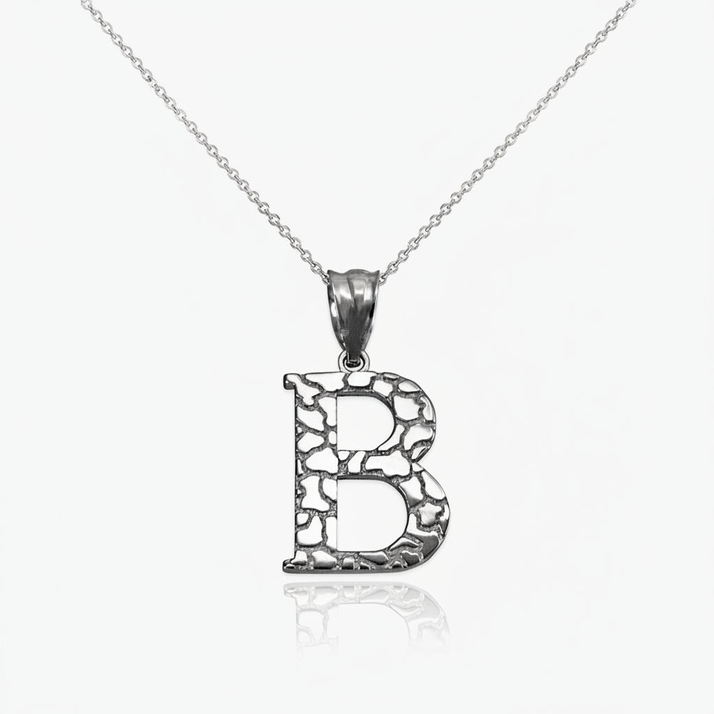 A silver initial pendant necklace featuring the letter 'B' with a textured, openwork design, suspended from a delicate silver chain against a white background with a soft reflection.