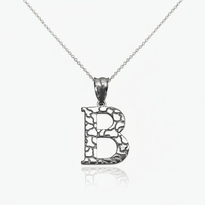 A silver initial pendant necklace featuring the letter 'B' with a textured, openwork design, suspended from a delicate silver chain against a white background with a soft reflection.