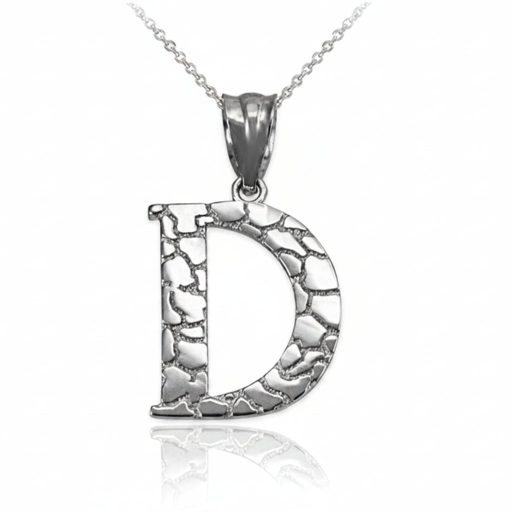A silver initial pendant necklace featuring the letter 'D' with a textured, hammered finish, suspended from a delicate silver chain on a white background with a soft reflection below.