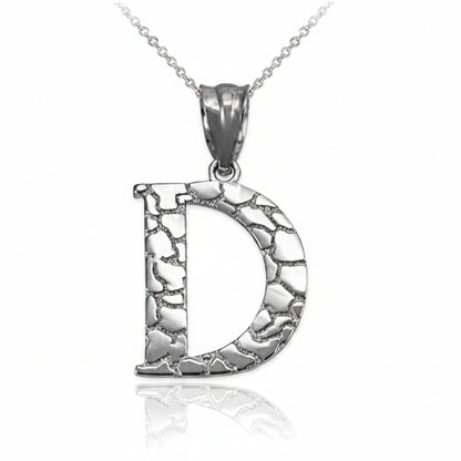 A silver initial pendant necklace featuring the letter 'D' with a textured, hammered finish, suspended from a delicate silver chain on a white background with a soft reflection below.