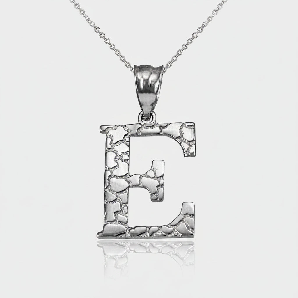 A silver initial pendant necklace featuring the letter 'E' with a textured, hammered finish and a polished bail, suspended from a delicate silver chain on a white background with a soft reflection.