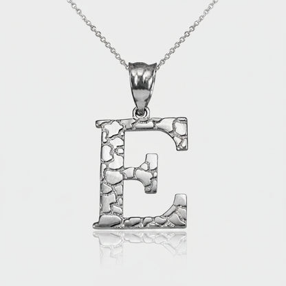 A silver initial pendant necklace featuring the letter 'E' with a textured, hammered finish and a polished bail, suspended from a delicate silver chain on a white background with a soft reflection.