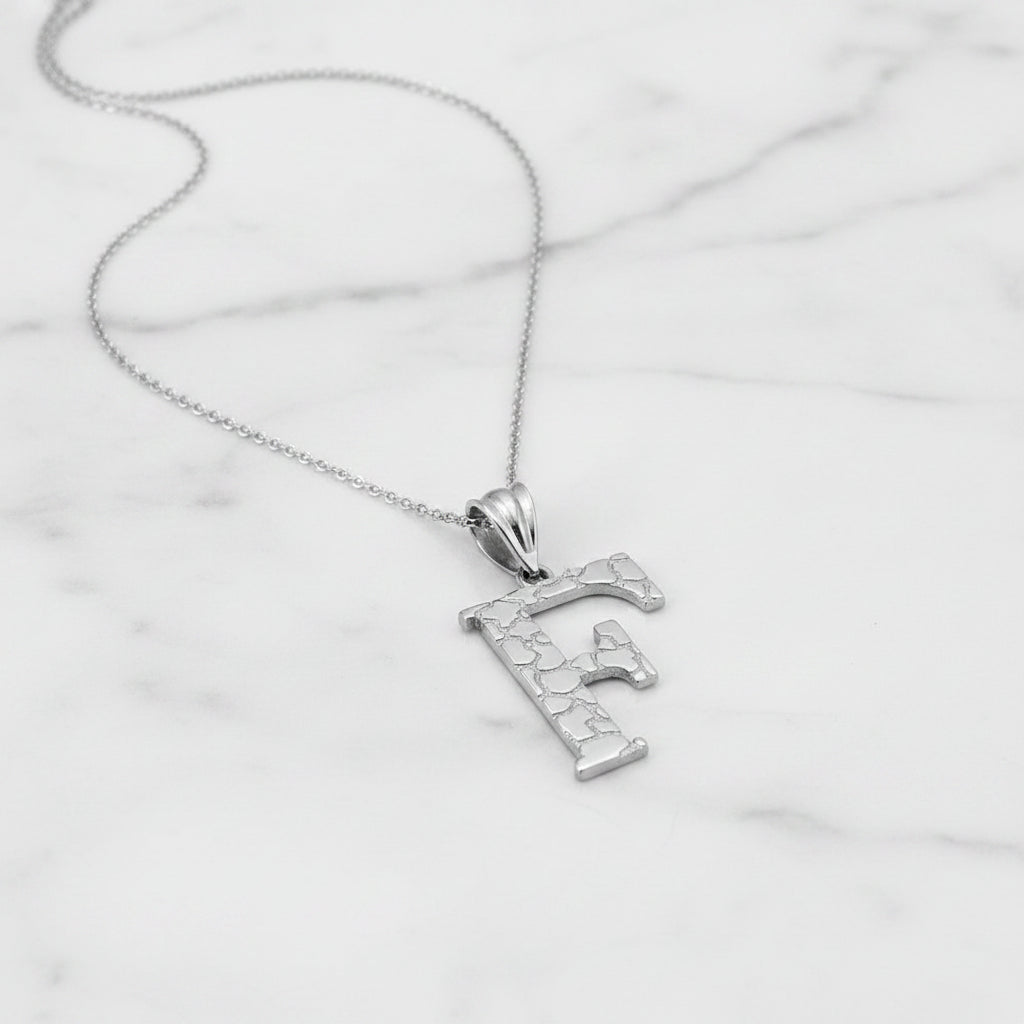 A silver initial pendant necklace featuring the letter 'F' with a textured, crackled finish, suspended from a delicate silver chain against a white background.