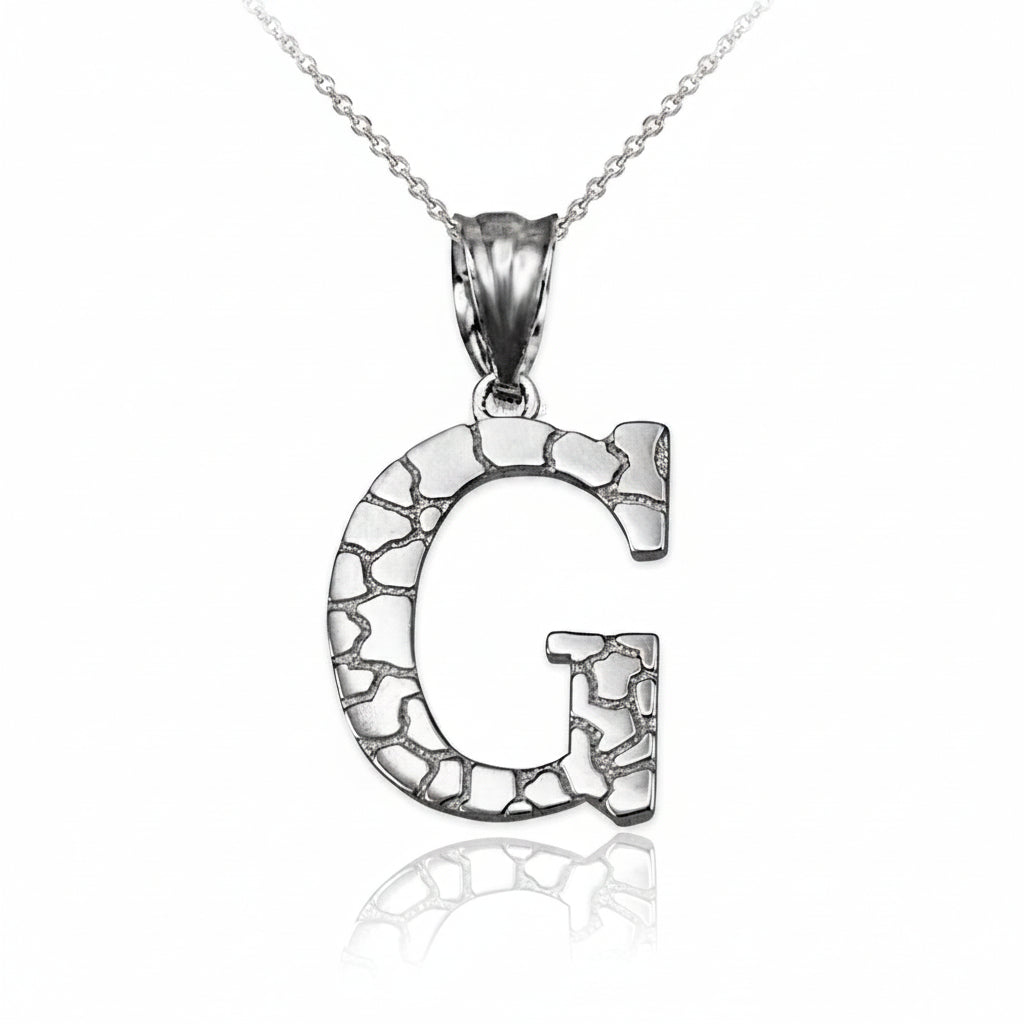 A silver initial pendant necklace featuring the letter 'G' with a textured, cracked stone-like pattern, suspended from a delicate silver chain on a white background with a soft reflection below.