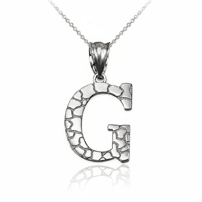 A silver initial pendant necklace featuring the letter 'G' with a textured, cracked stone-like pattern, suspended from a delicate silver chain on a white background with a soft reflection below.