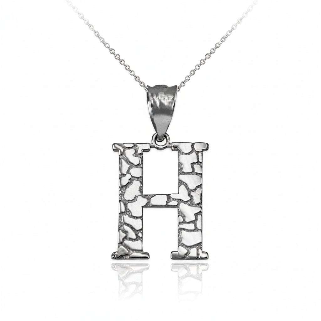 A sterling silver initial pendant necklace featuring the letter 'H' with a textured, openwork stone-like pattern, suspended from a delicate chain on a white background with a soft reflection.
