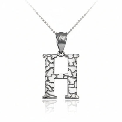 A sterling silver initial pendant necklace featuring the letter 'H' with a textured, openwork stone-like pattern, suspended from a delicate chain on a white background with a soft reflection.