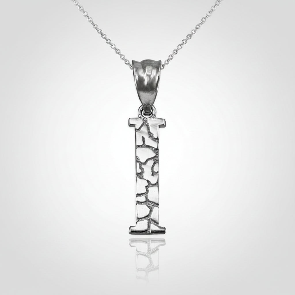 A silver initial pendant necklace featuring the letter 'I' with a textured, openwork design, suspended from a delicate silver chain on a white background with a soft reflection.