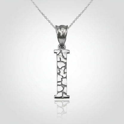 A silver initial pendant necklace featuring the letter 'I' with a textured, openwork design, suspended from a delicate silver chain on a white background with a soft reflection.