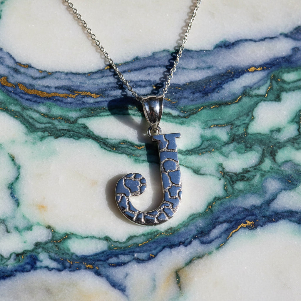 A sterling silver initial pendant necklace featuring the letter 'J' with a textured, openwork design, suspended from a delicate chain on a white background with a soft reflection below.