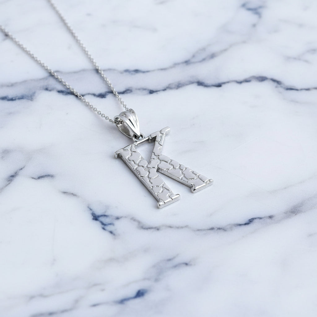 A silver initial pendant necklace featuring the letter 'K' with a textured, openwork design, suspended from a delicate silver chain on a white background with a soft reflection below.
