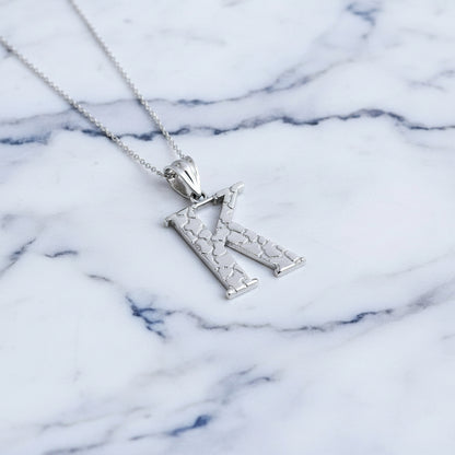 A silver initial pendant necklace featuring the letter 'K' with a textured, openwork design, suspended from a delicate silver chain on a white background with a soft reflection below.