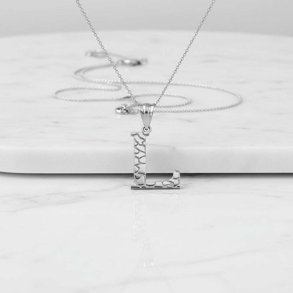 A silver initial pendant necklace featuring the letter 'L' with a textured, mosaic-like pattern on its vertical and horizontal bars, suspended from a delicate link chain against a white background with a soft reflection below.
