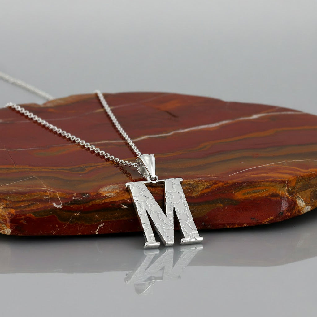 A silver initial pendant necklace featuring the letter 'M' with a textured, crackled pattern on a clean white background.