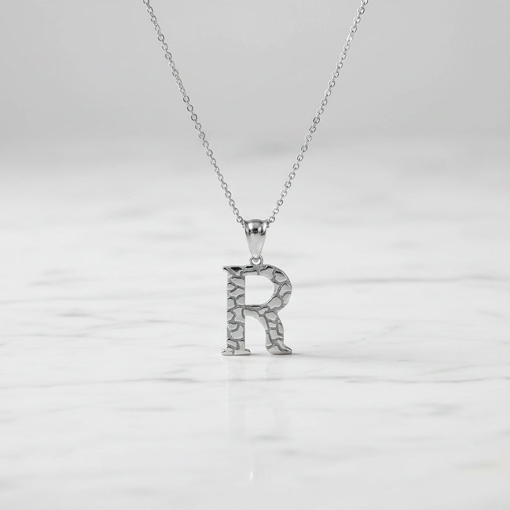 A silver initial pendant necklace featuring the letter 'R' with a textured, crackled surface design, suspended from a delicate silver chain against a white background.