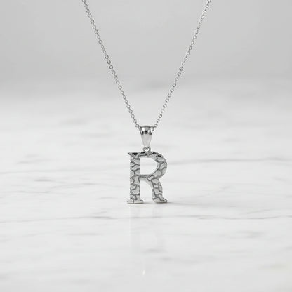 A silver initial pendant necklace featuring the letter 'R' with a textured, crackled surface design, suspended from a delicate silver chain against a white background.