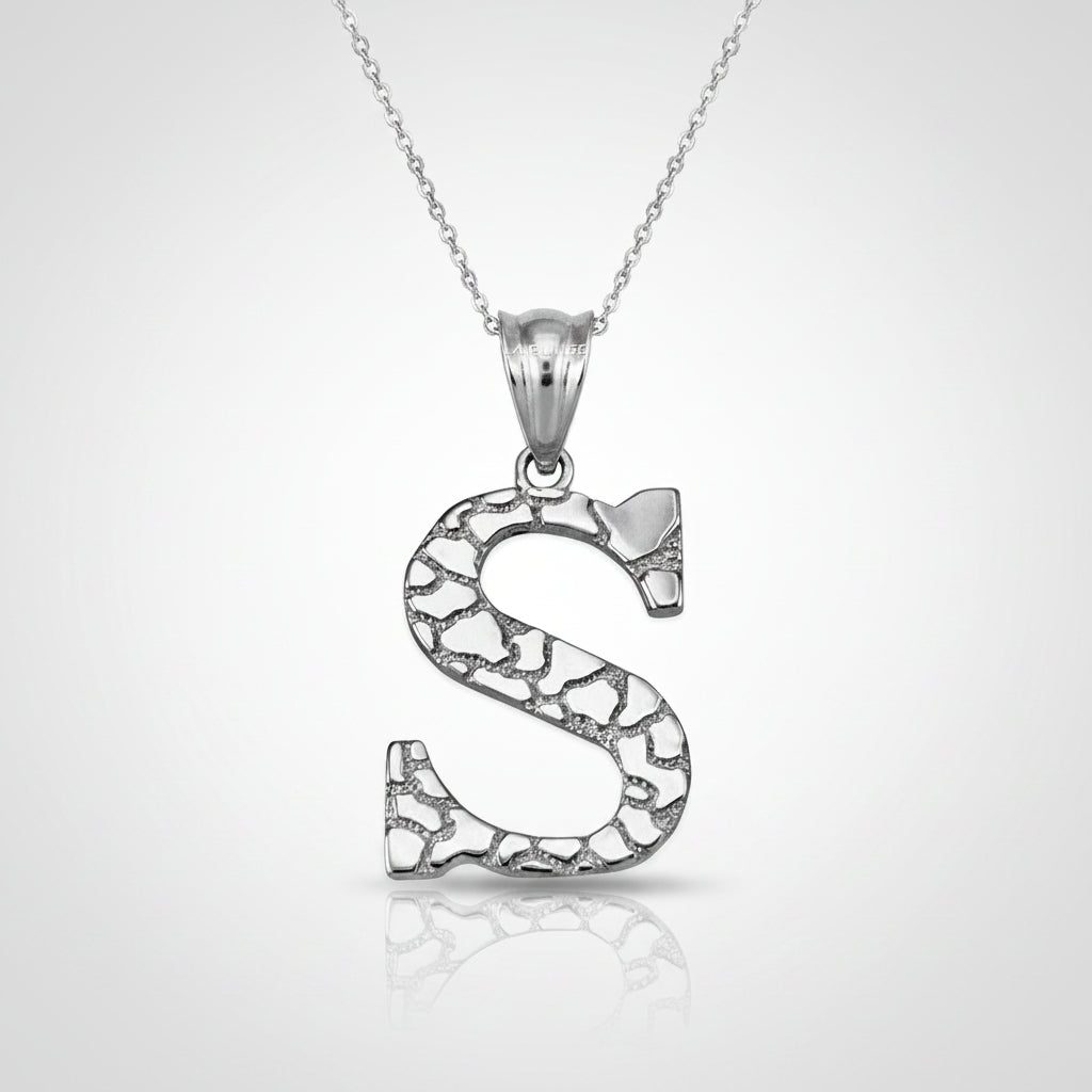 A silver initial pendant necklace featuring the letter 'S' with an openwork, textured mosaic pattern, suspended from a delicate silver chain on a white background with a soft reflection.