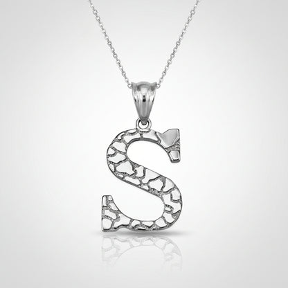 A silver initial pendant necklace featuring the letter 'S' with an openwork, textured mosaic pattern, suspended from a delicate silver chain on a white background with a soft reflection.