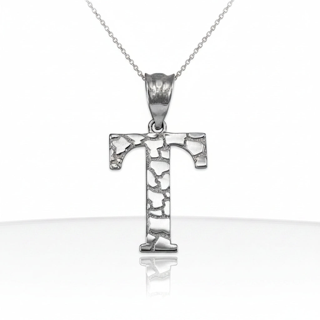 A silver initial pendant necklace featuring the letter 'T' with a textured, hammered finish, suspended from a delicate silver chain on a white background with a soft reflection below.