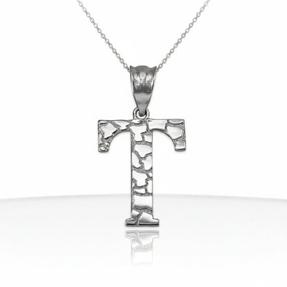 A silver initial pendant necklace featuring the letter 'T' with a textured, hammered finish, suspended from a delicate silver chain on a white background with a soft reflection below.