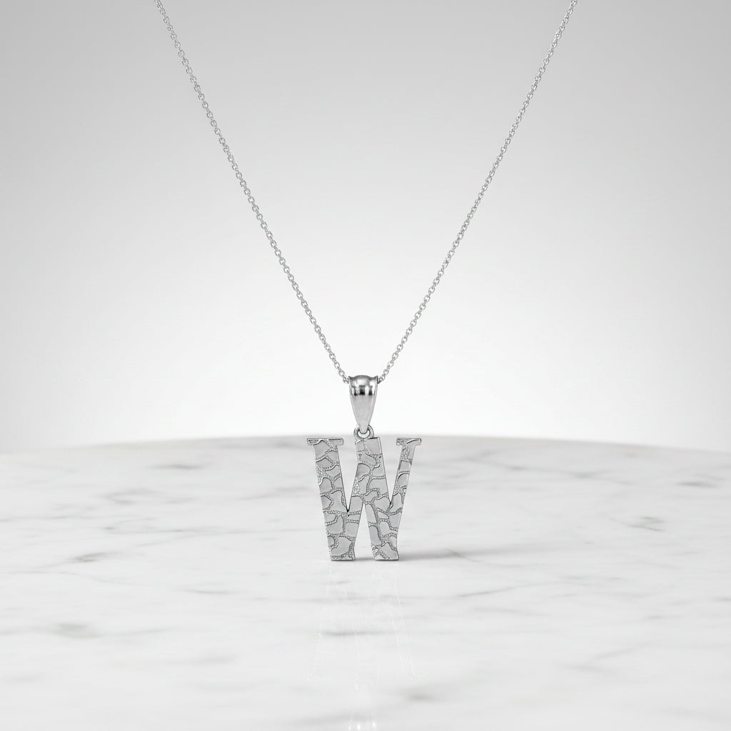 A silver initial pendant necklace featuring the letter 'W' with a textured, openwork geometric design, suspended from a delicate silver chain on a white background with a soft reflection.