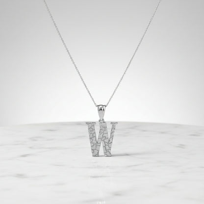 A silver initial pendant necklace featuring the letter 'W' with a textured, openwork geometric design, suspended from a delicate silver chain on a white background with a soft reflection.