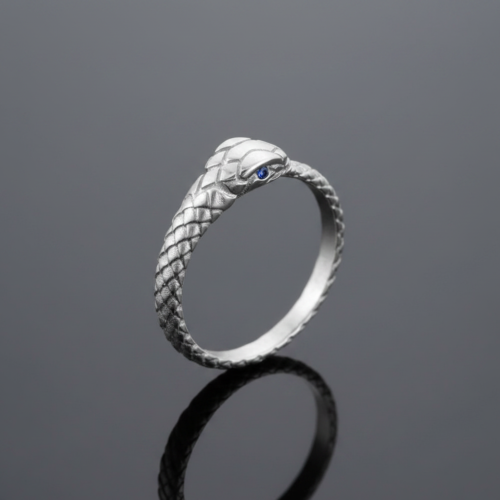 A silver snake ring with a textured scale pattern and a small blue gemstone eye, standing on a reflective dark gray surface.