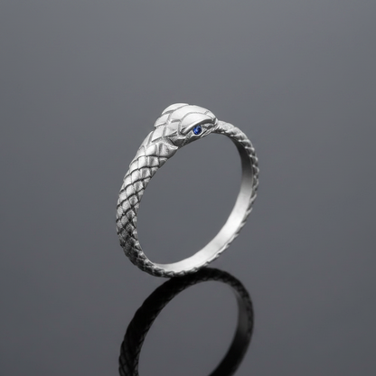 A silver snake ring with a textured scale pattern and a small blue gemstone eye, standing on a reflective dark gray surface.