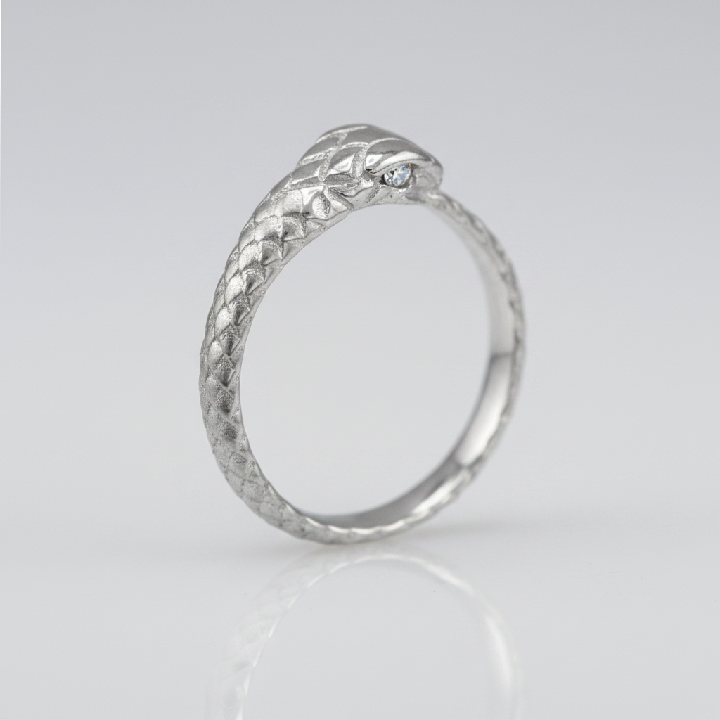 A silver snake ring with a textured scale pattern and a small clear gemstone in one eye, shown on a light gray reflective background.