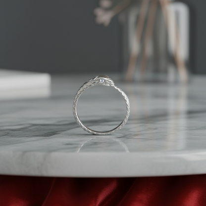 A silver snake ring with a small clear gemstone on its head, resting on a white marble surface with a blurred gray background.