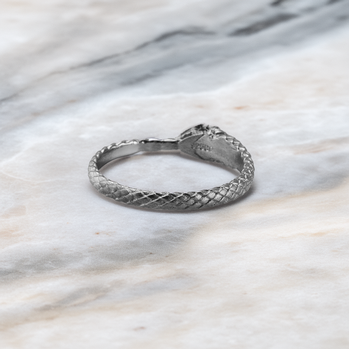 A silver snake ring with a textured, woven scale pattern, resting on a light gray and white marble surface.