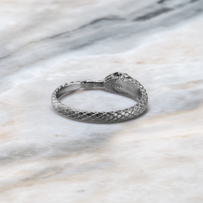 A silver snake ring with a textured, woven scale pattern, resting on a light gray and white marble surface.