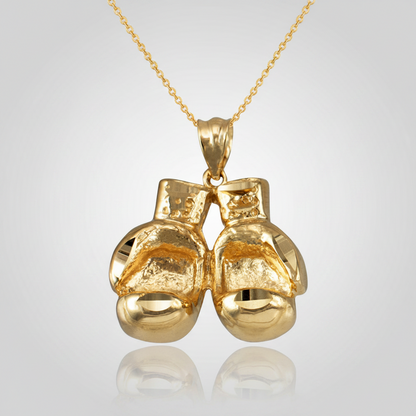 A gold pendant necklace featuring a pair of small, textured boxing gloves hanging from a delicate gold chain, set against a light gray gradient background with a soft reflection below.