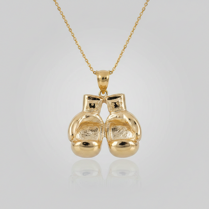 A gold pendant necklace featuring a pair of small boxing gloves hanging from a delicate gold chain, set against a light gray gradient background with a soft reflection below.