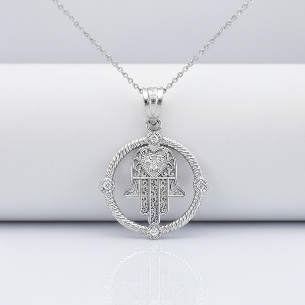 A silver Hamsa hand pendant necklace with a heart-shaped center and a rope-style circular border, displayed on a white cylindrical stand against a light gray background.