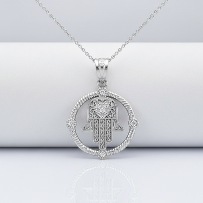 A silver Hamsa hand pendant necklace with a heart-shaped center and a rope-style circular border, displayed on a white cylindrical stand against a light gray background.