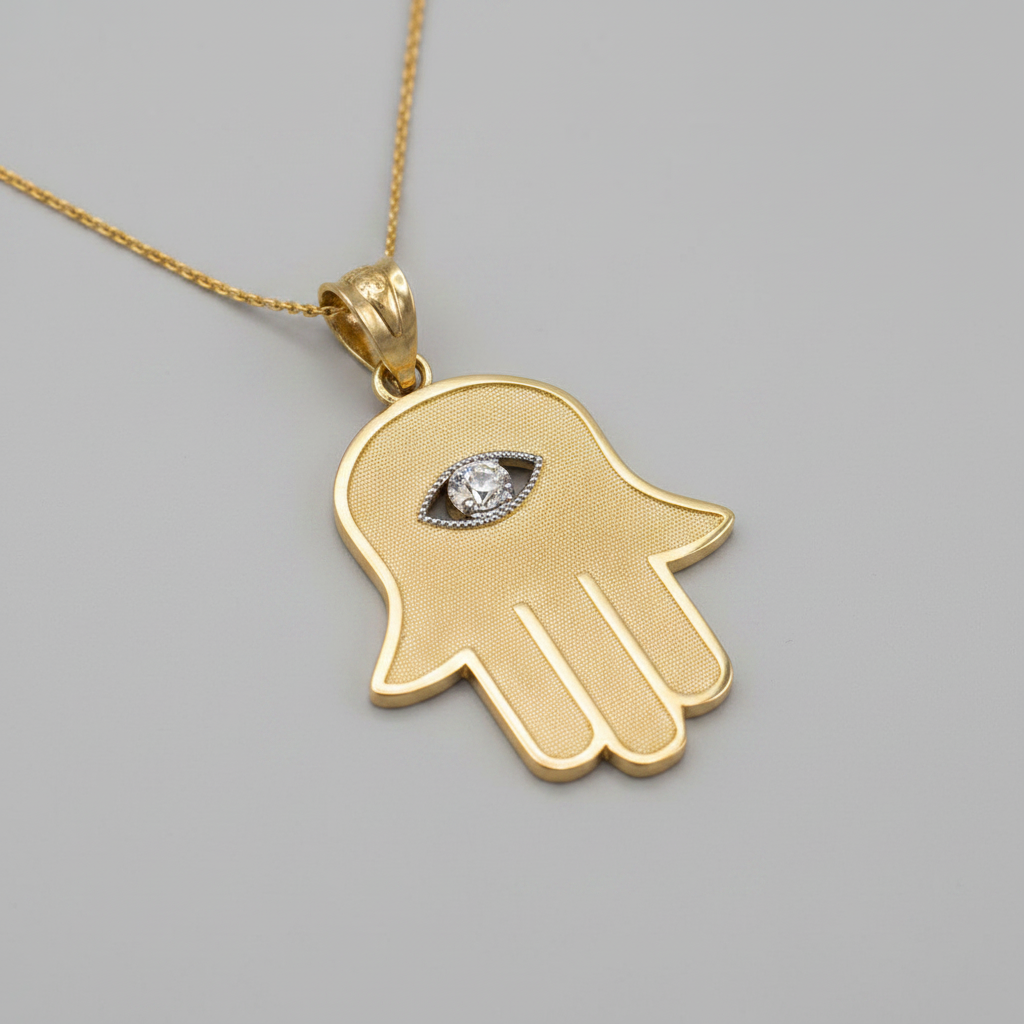 A gold Hamsa hand pendant necklace with an evil eye design in the center, featuring a textured finish and a small clear gemstone, shown on a plain gray background.