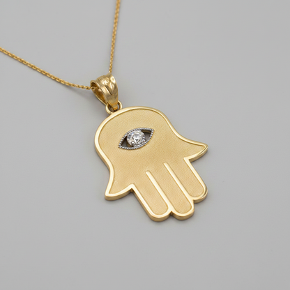 A gold Hamsa hand pendant necklace with an evil eye design in the center, featuring a textured finish and a small clear gemstone, shown on a plain gray background.