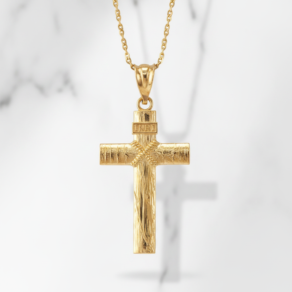 A gold cross pendant necklace with a textured finish and a 'INRI' inscription on the top, hanging from a delicate gold chain against a light gray marble background.