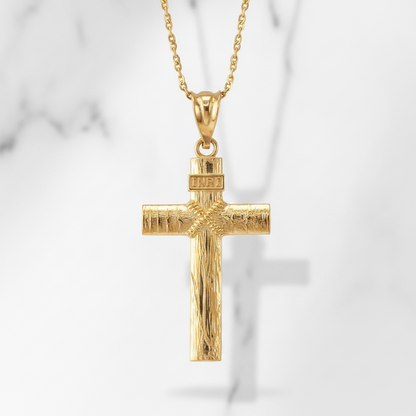 A gold cross pendant necklace with a textured finish and a 'INRI' inscription on the top, hanging from a delicate gold chain against a light gray marble background.