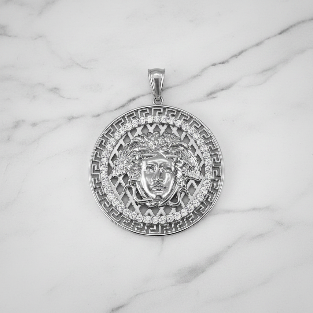 A round silver pendant featuring a Medusa head design, surrounded by Greek key patterns and small clear stones, displayed on a white marble background.