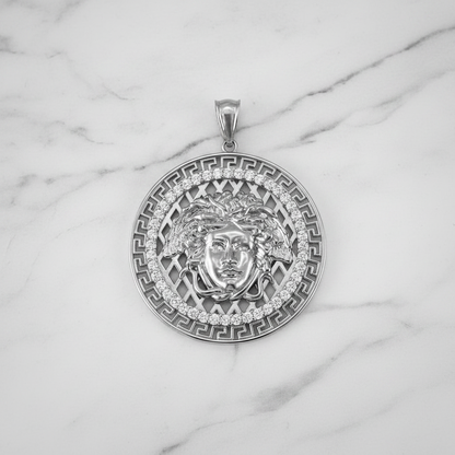 A round silver pendant featuring a Medusa head design, surrounded by Greek key patterns and small clear stones, displayed on a white marble background.