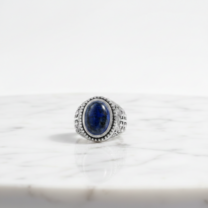 A silver-toned signet ring with an oval dark blue lapis lazuli stone, set on a white marble surface against a white background.