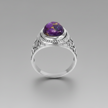 A silver ring with an ornate filigree band and a round purple gemstone featuring gold inclusions, shown on a neutral gray background.
