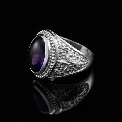 A silver-toned signet ring with an oval purple amethyst cabochon, featuring intricate filigree detailing on the band, set against a solid black background with a clear reflection below.