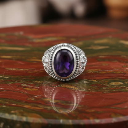 A silver-toned ring with an oval purple amethyst center stone and small clear accent stones, displayed on a reflective red and green marbled surface.
