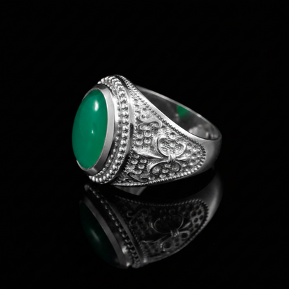 A silver-toned ring with an oval green cabochon gemstone and ornate engraved detailing on the band, displayed on a reflective black surface.