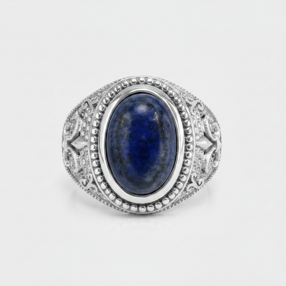 An oval silver ring featuring a deep blue lapis lazuli stone in a detailed filigree setting, photographed on a light gray background.