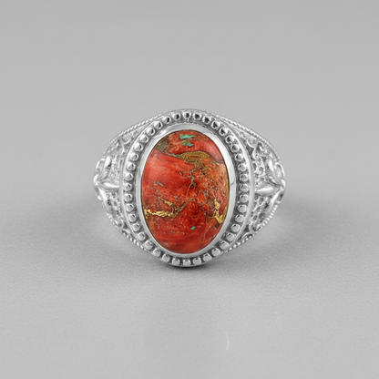A silver ring featuring an oval red jasper stone with prominent gold and green matrix patterns, set in an ornate filigree-style silver band on a neutral gray background.