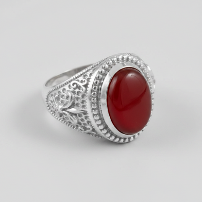 A silver signet ring featuring a large, smooth, oval-shaped deep red cabochon gemstone, set in an ornate filigree band with beaded detailing, shown on a neutral light gray background.